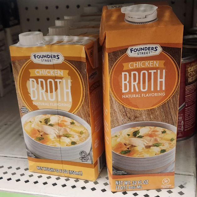 Two cartons of Founders Street Chicken Broth with natural flavoring are displayed on a store shelf. Both cartons feature an image of soup and text indicating a 32-ounce (946 ml) quantity—no need to pay more for less.