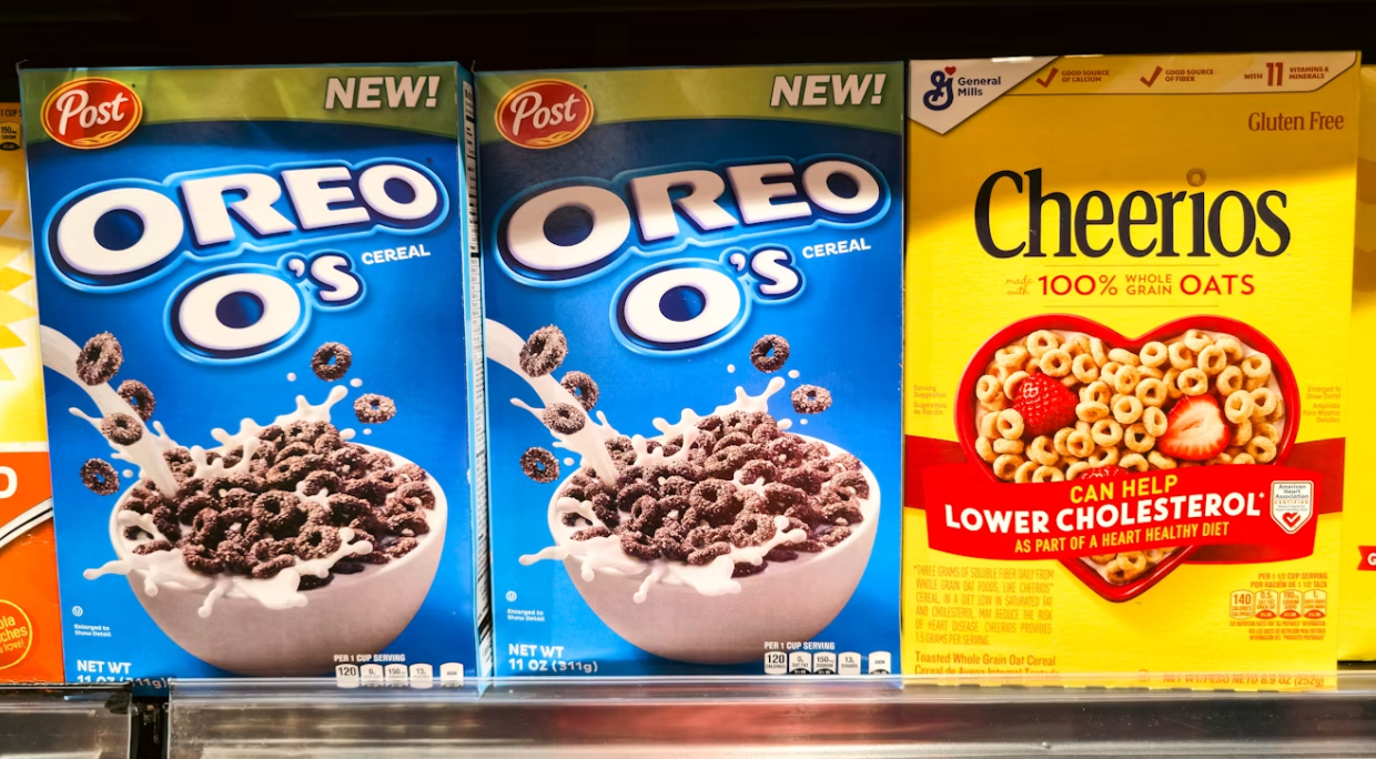 Two boxes of Oreo O’s cereal sit on a shelf next to Cheerios, all brightly lit with colorful packaging—reminding shoppers not to pay more for less when choosing their favorite cereals.