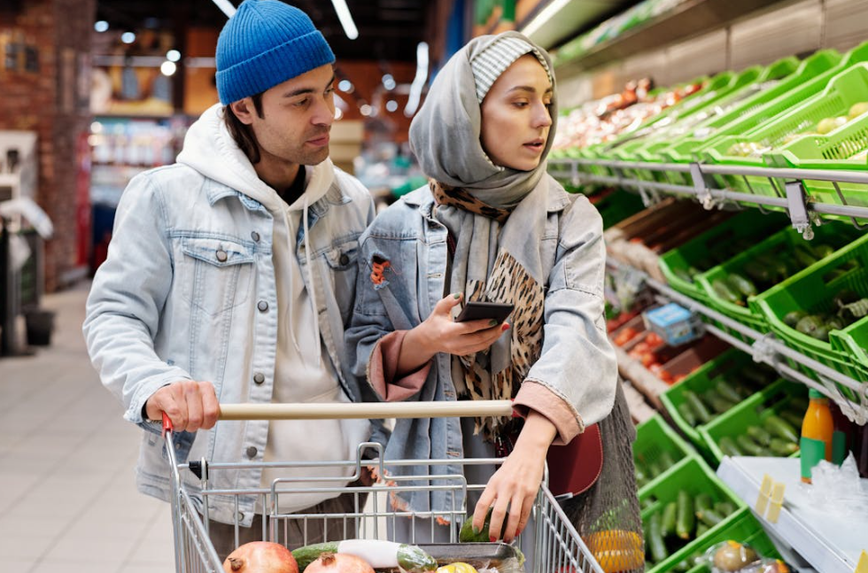 A man and woman shop for groceries together, pushing a cart filled with produce. Despite having to pay more for less, the woman in a headscarf checks her phone and reaches for vegetables while the man stands beside her in a blue beanie and denim jacket.