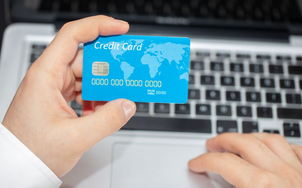 A person holding a blue credit card above a laptop keyboard, preparing for an online transaction. The card features a world map design and EMV chip, reminding us not to pay more for less when shopping online.