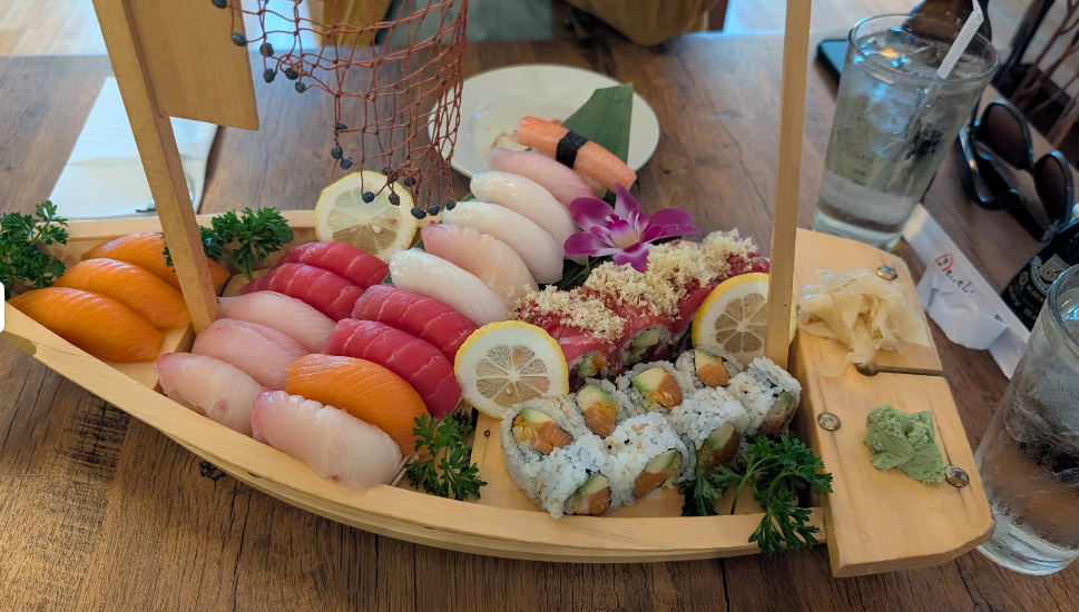 A wooden sushi boat holds various sushi pieces, including nigiri and rolls, garnished with lemon slices, wasabi, pickled ginger, parsley, and a purple orchid, set on a wooden table with drinks and utensils nearby.