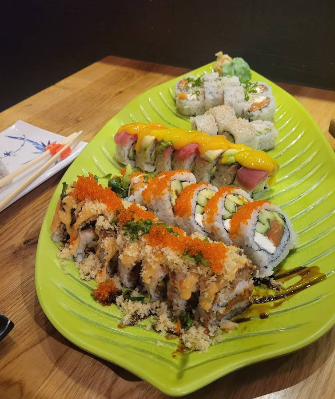 A green leaf-shaped plate filled with assorted sushi rolls, topped with colorful sauces, fish roe, and garnishes, sits on a wooden table next to chopsticks and a small dish.