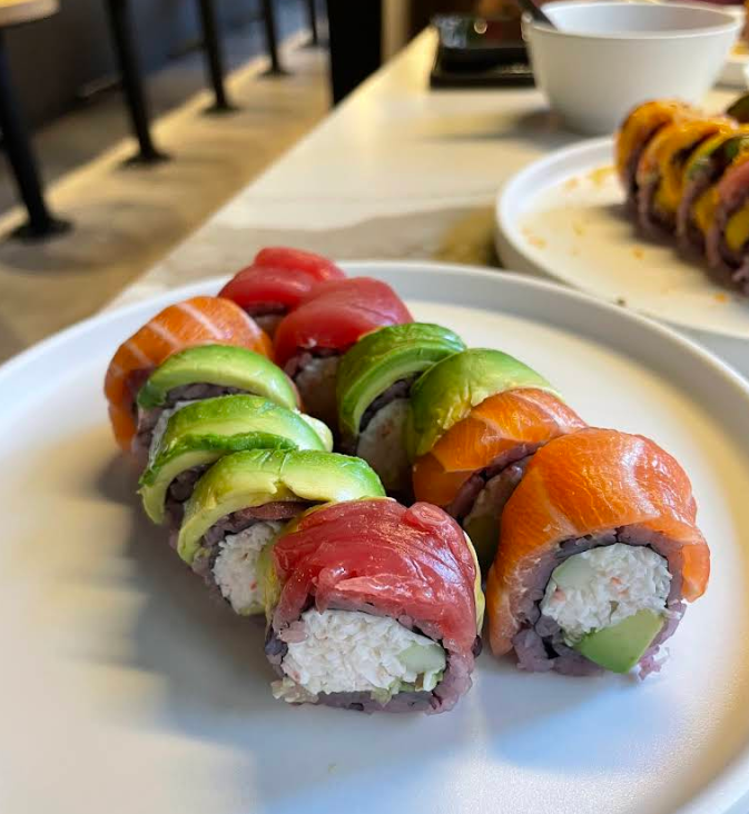 A plate of colorful sushi rolls topped with slices of salmon, tuna, and avocado, filled with crab and cucumber, sits on a white plate in a modern restaurant setting. Another sushi roll is visible in the background.