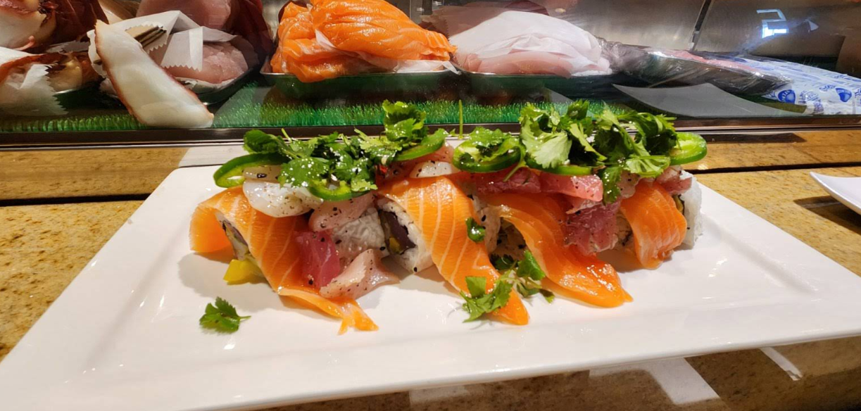 A close-up of two sushi rolls topped with slices of salmon, tuna, jalapeño, cilantro, and sesame seeds, served on a white rectangular plate at a sushi bar. Assorted fish are displayed in the background.