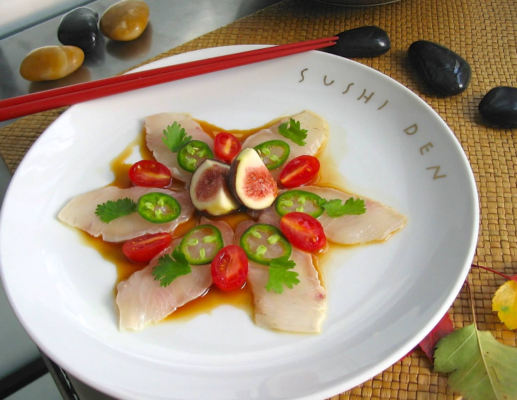 A plate of sashimi is artfully arranged with white fish slices, cherry tomatoes, jalapeño slices, cilantro, and fig halves on soy sauce, garnished with red chopsticks and stones nearby. The plate is labeled "Sushi Den.