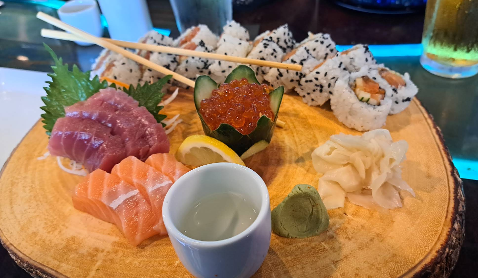 A wooden platter with sliced raw tuna and salmon, sushi rolls with sesame seeds, a cucumber cup filled with salmon roe, pickled ginger, wasabi, a lemon slice, and a small cup of sake.