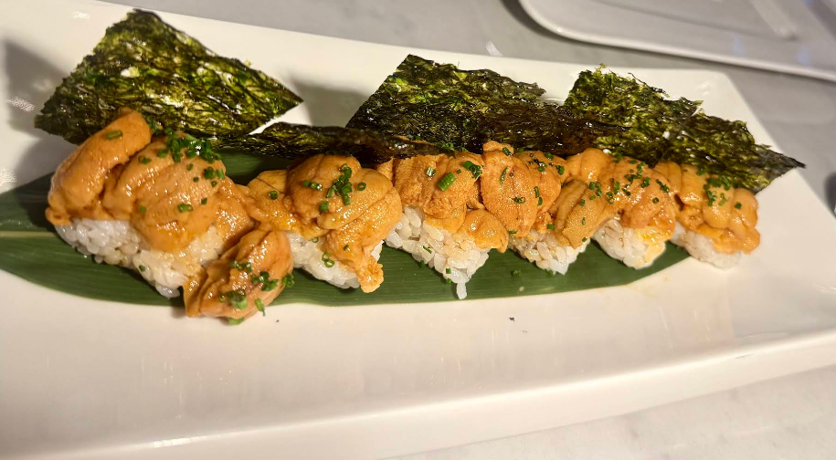 A row of sushi pieces topped with fresh orange uni (sea urchin), garnished with chopped chives and served on a green leaf, each piece accompanied by a rectangular sheet of nori on a white plate.