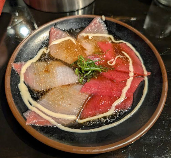 A black plate with thin slices of raw fish sashimi, arranged in a circular pattern, garnished with a swirl of sauce and topped with chopped green onions, served in a shallow pool of soy-based sauce.
