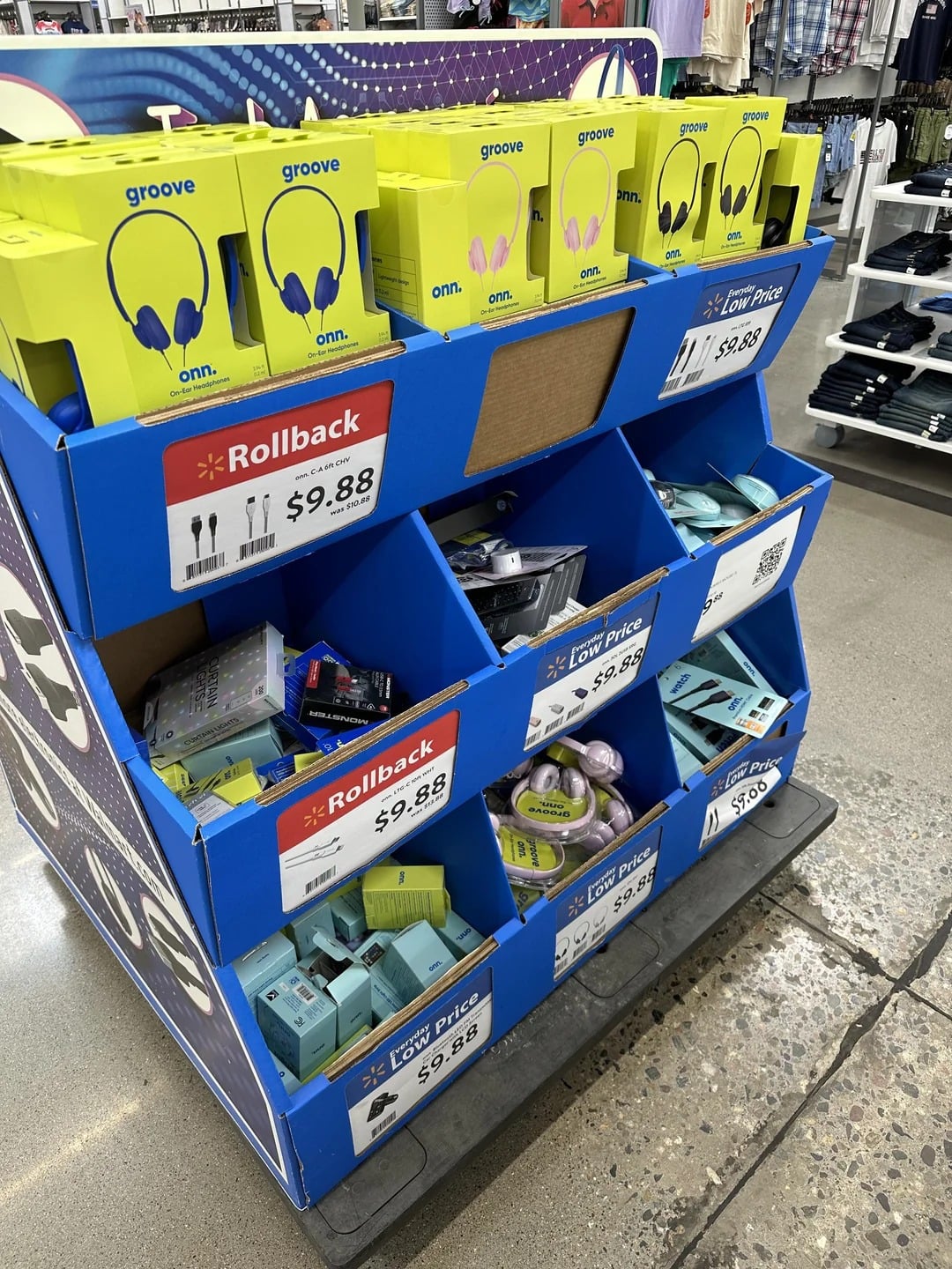 A blue store display holds yellow and green boxes of Walmart products like headphones, electronic accessories, and phone chargers. Rollback price signs show items are $9.88 or $7.96, with clothing racks visible in the background.