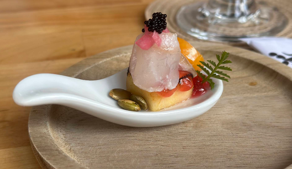 A white ceramic spoon holds a gourmet appetizer with a slice of raw fish, black caviar, pickled onions, berries, a yellow base, pumpkin seeds, and a small fern garnish, all on a wooden plate.