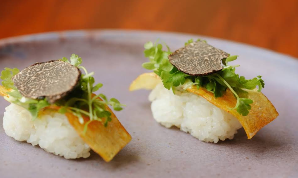 Two pieces of elegant sushi topped with microgreens, a thin slice of black truffle, and a crisp yellow chip, presented on a smooth ceramic plate.