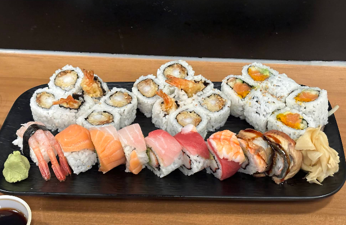 A black tray with assorted sushi, including shrimp, tuna, salmon, eel, and various sushi rolls. Pickled ginger, wasabi, and a dish of soy sauce are on the side.