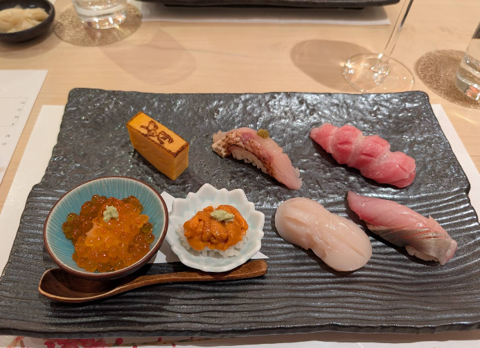 A rectangular black plate with assorted sushi pieces, including tamago, nigiri with various fish, scallop, sea urchin, and salmon roe in small bowls, served with a wooden spoon. Wine glasses and water are visible in the background.