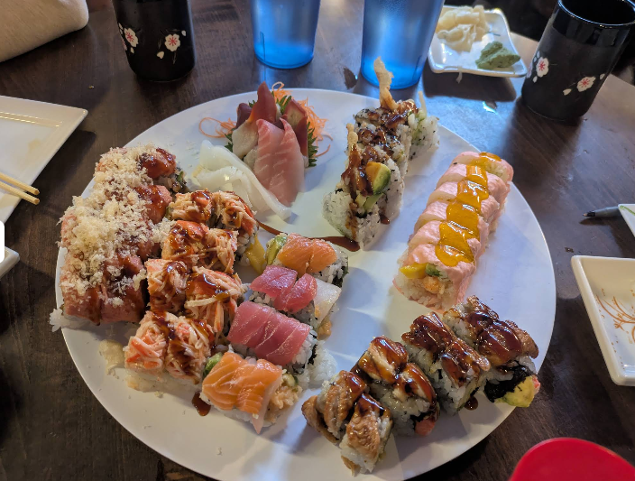 A large white plate filled with assorted sushi rolls, nigiri, and sashimi, with various colorful toppings and sauces, sits on a wooden table with drinks, soy sauce, chopsticks, and condiments in the background.