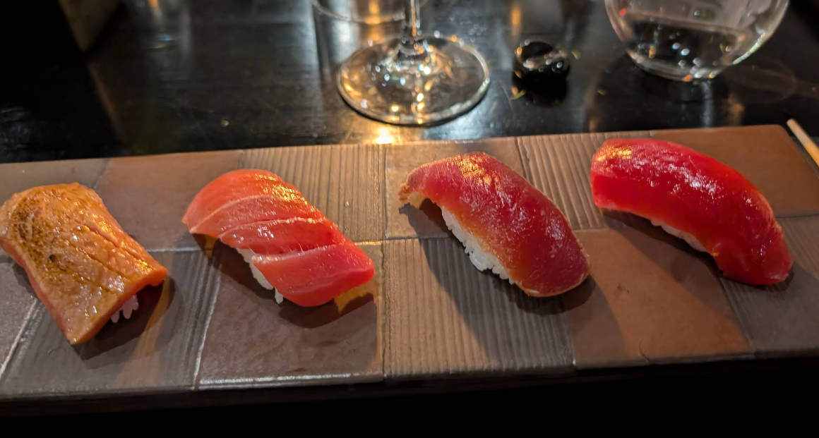 A rectangular plate with four pieces of sushi, each topped with different types of raw fish, sits on a dark table. A wine glass and drinking glass are visible in the background.