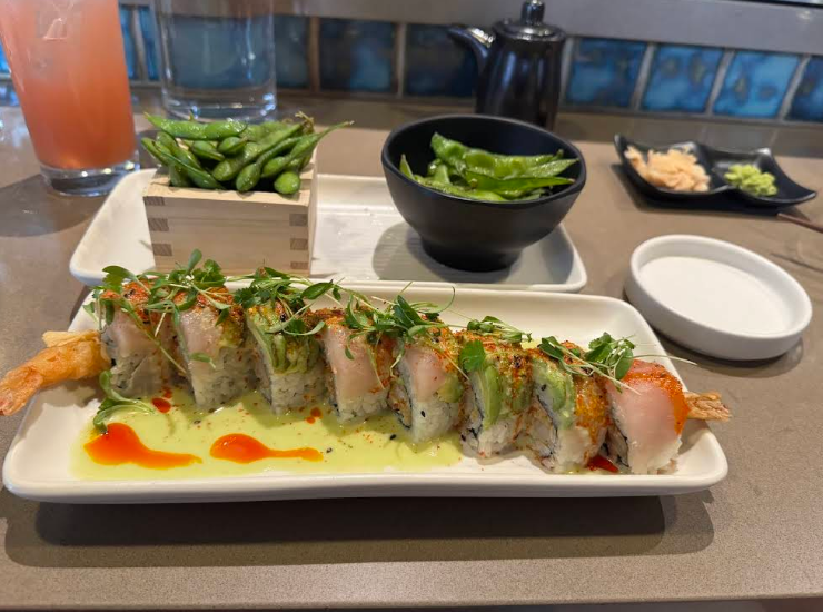 A plate of sushi rolls topped with greens and sauce, with shrimp tempura inside, sits in front of edamame, a glass of pink drink, soy sauce, and small dishes of ginger and wasabi on a restaurant table.