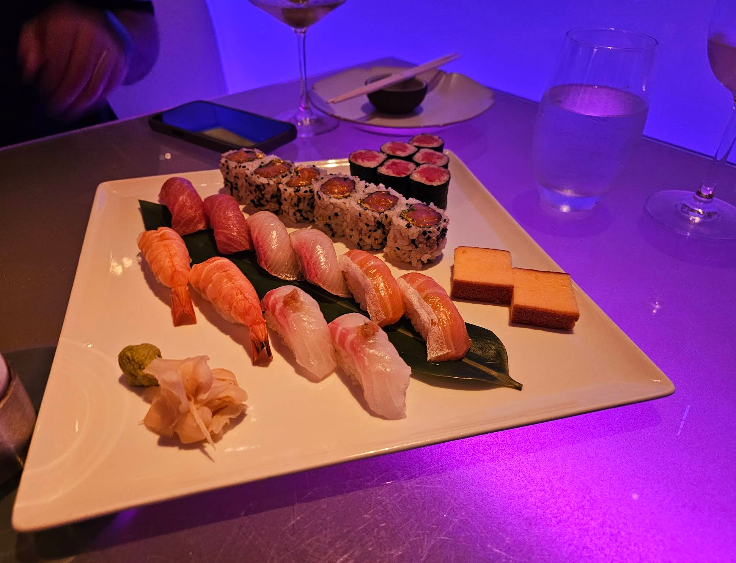 A square white plate with assorted sushi, including nigiri, maki rolls, and tamago slices, garnished with wasabi and pickled ginger, on a reflective table with drinks and a soft purple light.