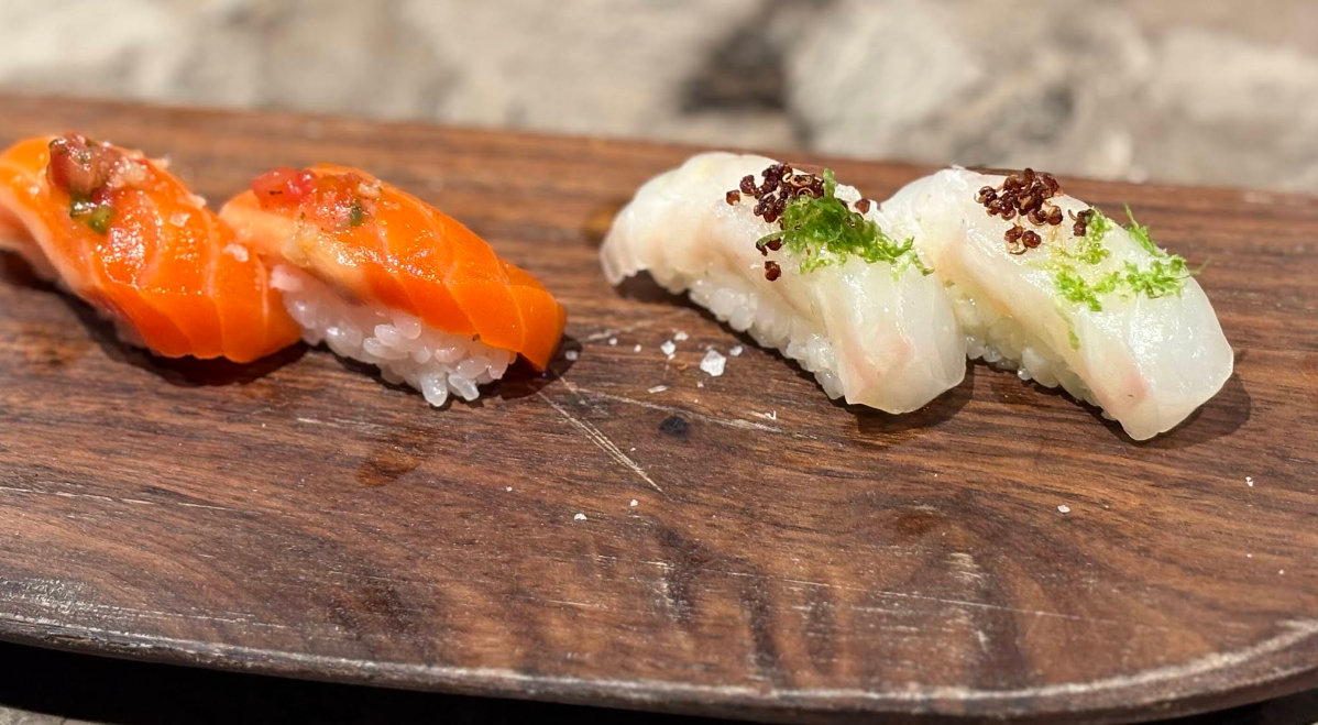 Two pieces of salmon nigiri sushi and two pieces of white fish nigiri sushi, each topped with garnishes, arranged on a wooden serving board.