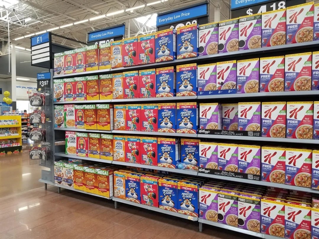 Store aisle with shelves stocked full of various breakfast cereals in colorful boxes, including Cheerios, Frosted Flakes, Froot Loops, Raisin Bran, and Special K—classic Walmart products. Price tags are visible above and below the shelves.
