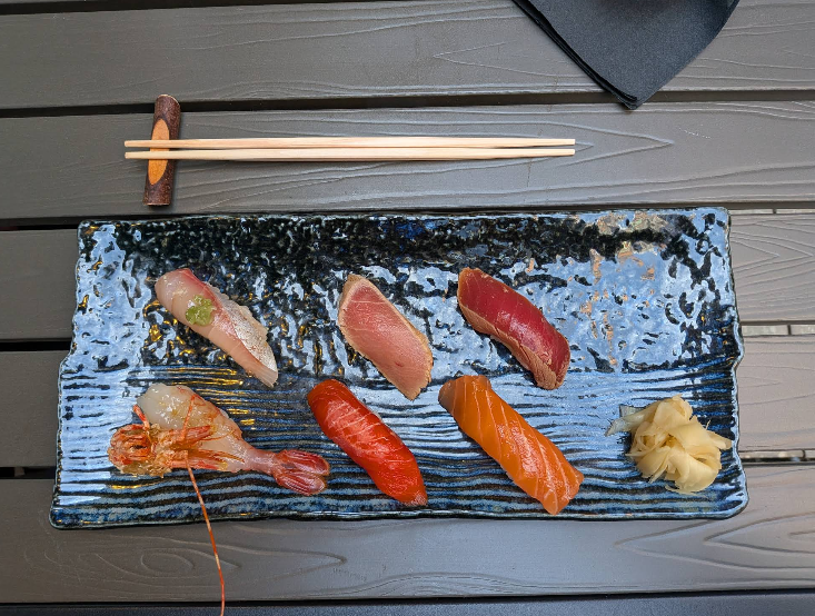 A rectangular dark ceramic plate with six pieces of assorted nigiri sushi, a small pile of pickled ginger, and a pair of chopsticks resting on a wooden holder, all on a wooden table.
