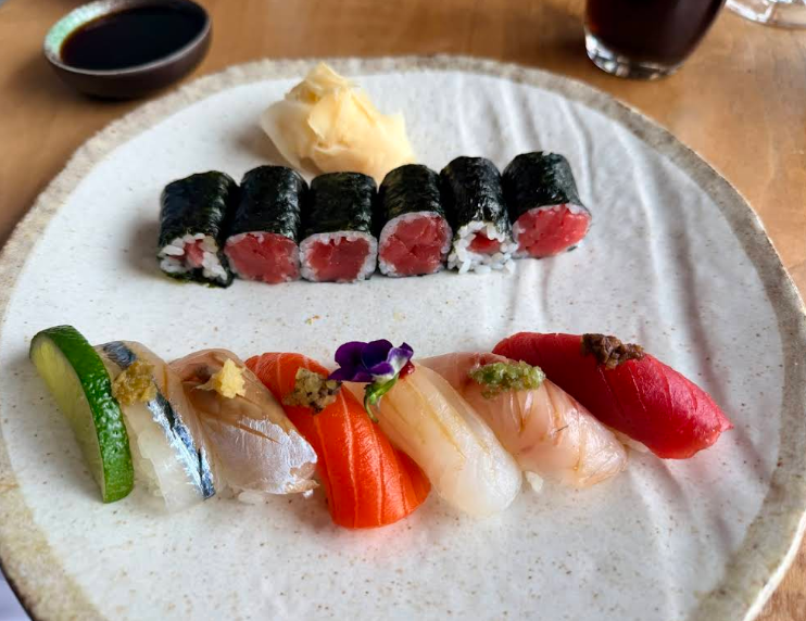 A plate of assorted sushi, including six tuna maki rolls, five pieces of nigiri with various fish and garnishes, a slice of cucumber, pickled ginger, and a small dish of soy sauce in the background.