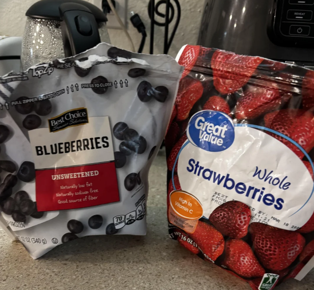 Two bags of frozen fruit on a kitchen counter—Best Choice unsweetened blueberries and Great Value whole strawberries—are perfect to buy in bulk. Appliances and a kettle can be seen in the background.