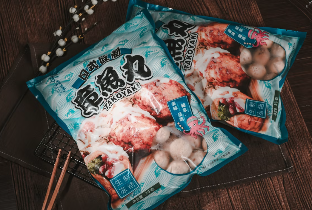 Two blue packages of frozen takoyaki balls with Japanese and Chinese text are placed on a dark wooden surface, next to chopsticks and decorative white flowers—perfect for those looking to buy in bulk for parties or gatherings.