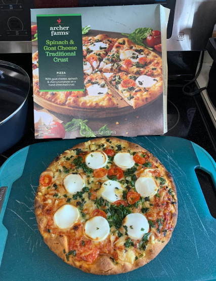 A cooked spinach and goat cheese pizza sits on a blue cutting board in front of its Archer Farms box, which shows the same pizza topped with goat cheese, spinach, and tomatoes—perfect to buy in bulk for easy meals at home.