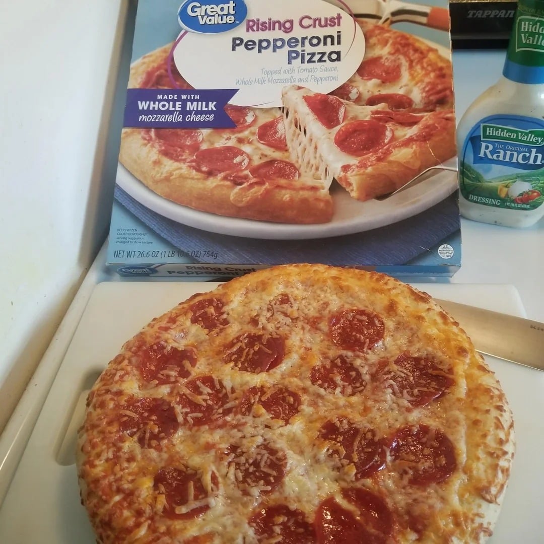 A cooked pepperoni pizza sits on a stovetop in front of its "Great Value Rising Crust Pepperoni Pizza" packaging, showcasing popular Walmart products. A bottle of Hidden Valley Ranch dressing is visible in the background.