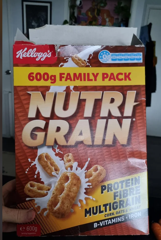 A hand holds a 600g family pack box of Kellogg's Nutri-Grain cereal, featuring splashing milk and cereal pieces on the front. The box highlights protein, fibre, multigrain, B-vitamins, and iron.