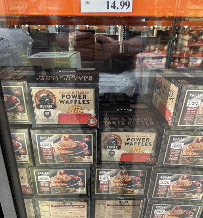 Boxes of Kodiak Protein-Packed Power Waffles are stacked in a freezer display at a store, with a price tag of $14.99 for those looking to buy in bulk. A reflection of the person taking the photo appears in the glass.