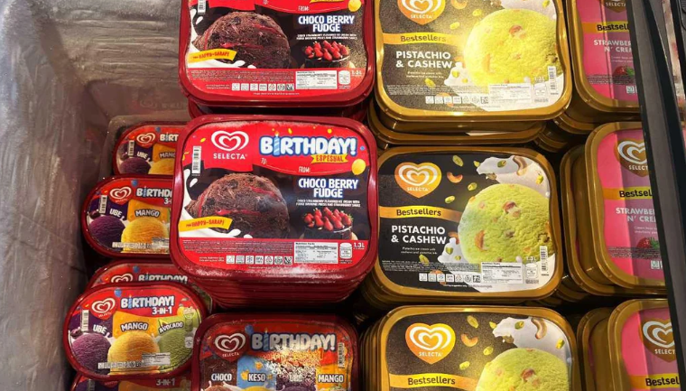 Ice cream tubs in a freezer, including red containers labeled "Birthday! Choco Berry Fudge" and yellow "Bestsellers Pistachio & Cashew." Smaller tubs with other flavors are also visible on the left—perfect if you want to buy in bulk.
