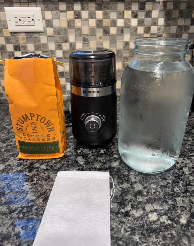 A bag of Stumptown Trapper Creek coffee, a black SHARDOR coffee grinder, a large mason jar filled with water, and a white paper coffee filter on a kitchen counter.