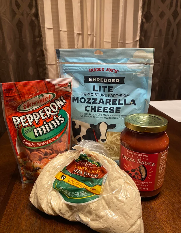 A bag of mini pepperoni slices, a bag of shredded mozzarella cheese, a jar of pizza sauce, and a ball of pizza dough are displayed on a wooden table with a striped curtain in the background.