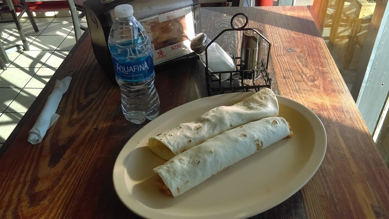 Two rolled tortillas on a beige plate—a classic find at top breakfast burrito spots—sit beside a bottle of Aquafina water on a wooden table. A napkin dispenser, metal condiment holder, and wrapped utensils rest in the sunlight from a nearby window.