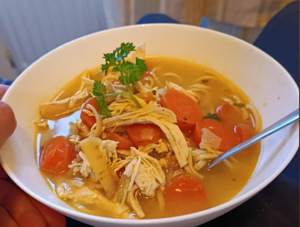 A bowl of chicken noodle soup with shredded chicken, sliced carrots, noodles, and fresh parsley on top. A spoon rests in the bowl, and a hand is holding the bowl.