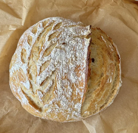 A round loaf of artisan bread with a golden crust, decorated with leaf-shaped scoring and dusted with flour, sits on a piece of brown parchment paper.