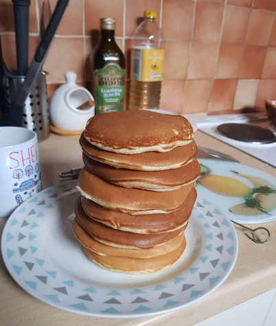 A tall stack of golden-brown pancakes sits on a patterned plate in a kitchen, with cooking utensils, a mug, and bottles of olive oil and vegetable oil visible in the background.