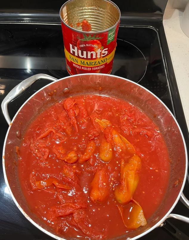 A pan filled with simmering whole peeled tomatoes in tomato sauce sits on a stovetop, perfect for cooking from scratch. An empty can labeled “Hunt’s San Marzano Whole Peeled Tomatoes” is placed behind the pan.
