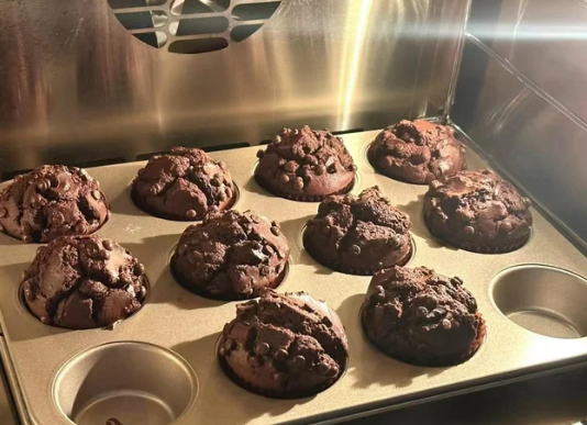 A tray of freshly baked chocolate muffins with chocolate chips, made from cooking from scratch, sits in an oven. Golden light illuminates the muffins and highlights their rich, textured tops, while one cup in the tray stands empty.