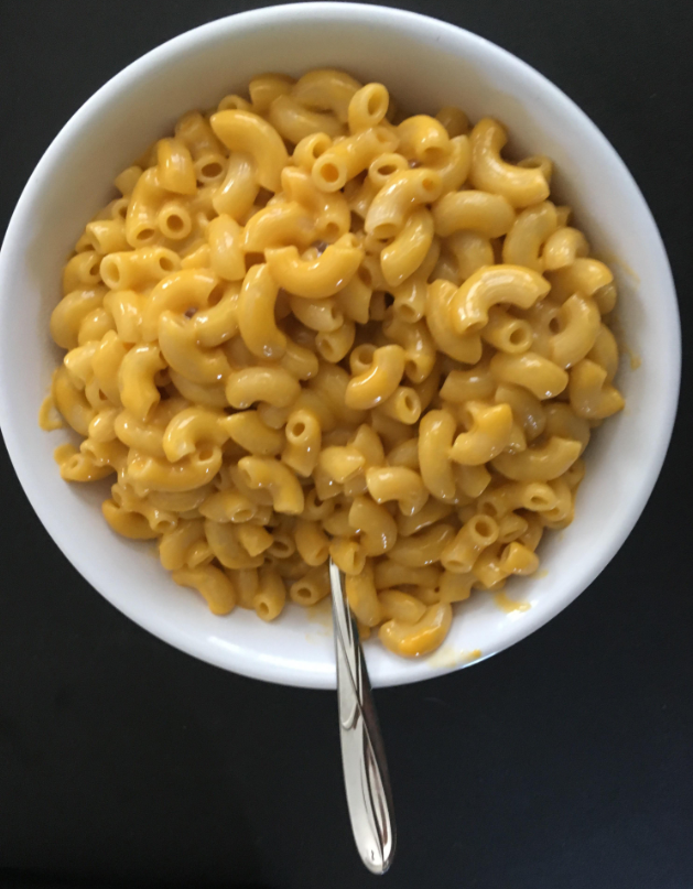 A white bowl filled with creamy macaroni and cheese, made by cooking from scratch, sits on a dark surface with a silver spoon resting inside the bowl.