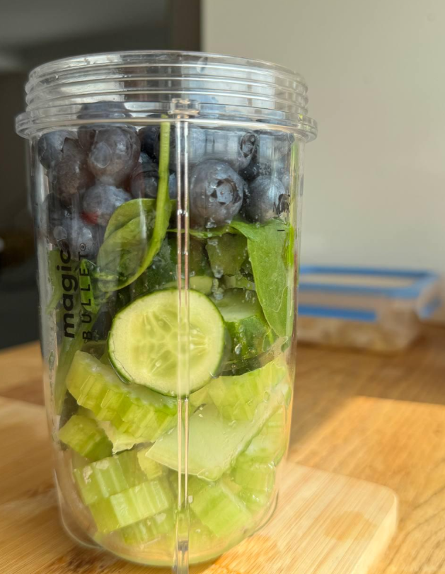 A clear blender cup filled with chopped celery, cucumber slices, spinach leaves, and blueberries sits on a wooden surface in bright sunlight—a perfect start for anyone who enjoys cooking from scratch.