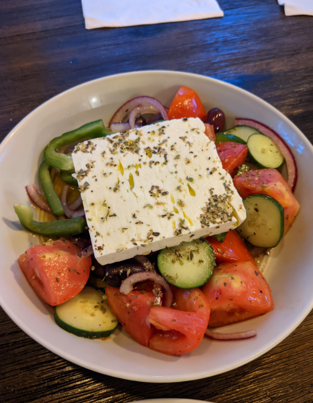 A bowl of Greek salad with chopped tomatoes, cucumbers, red onions, green peppers, olives, and a large slice of feta cheese topped with herbs, served on a wooden table—perfect for anyone who loves cooking from scratch.