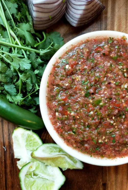 A bowl of fresh salsa made by cooking from scratch sits on a wooden surface, surrounded by cilantro, a jalapeño pepper, red onion pieces, and lime wedges.