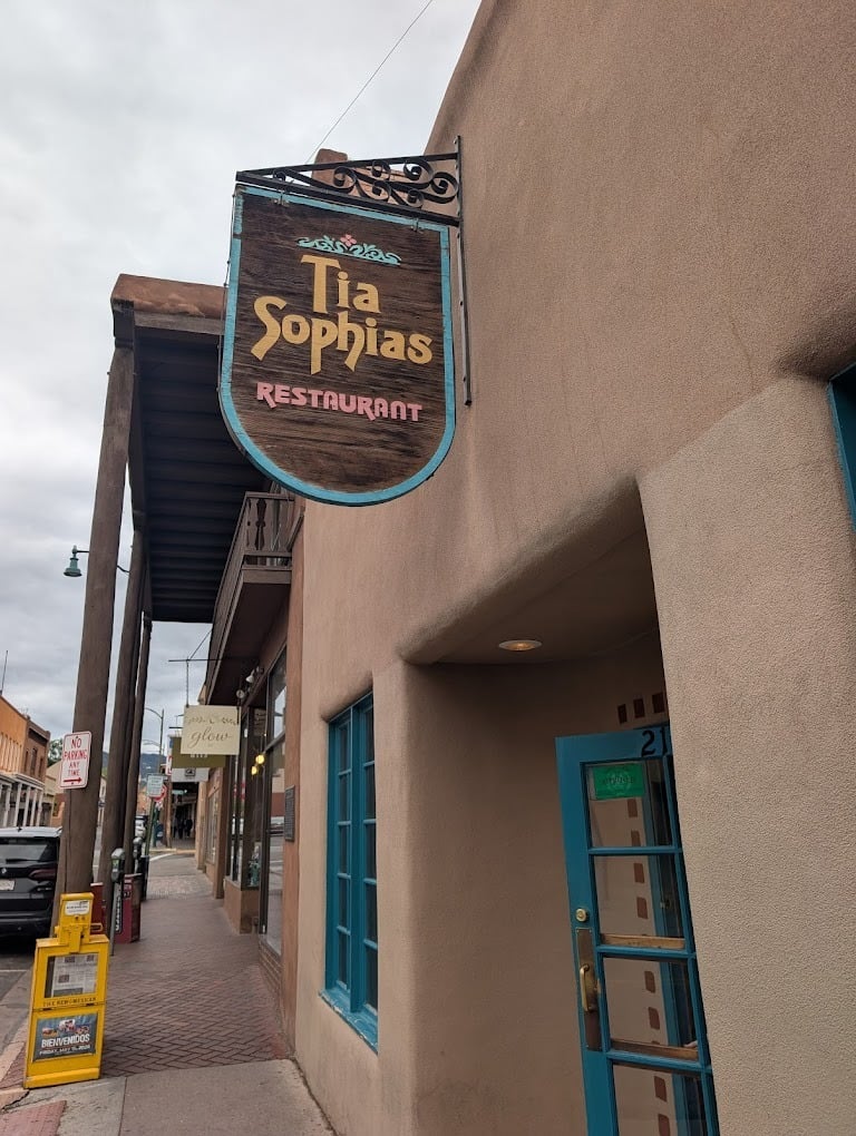 A wooden sign for “Tia Sophia’s Restaurant,” one of Santa Fe’s classic breakfast burrito spots, hangs above the entrance of an adobe-style building on a sidewalk lined with shops and newspaper stands. The sky is cloudy.