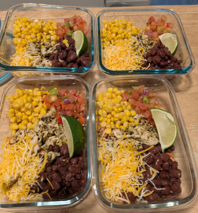 Four glass meal prep containers filled with shredded cheese, black beans, corn, pico de gallo, shredded chicken, and a lime wedge—perfect for those who love cooking from scratch and organizing healthy meals on a wooden surface.