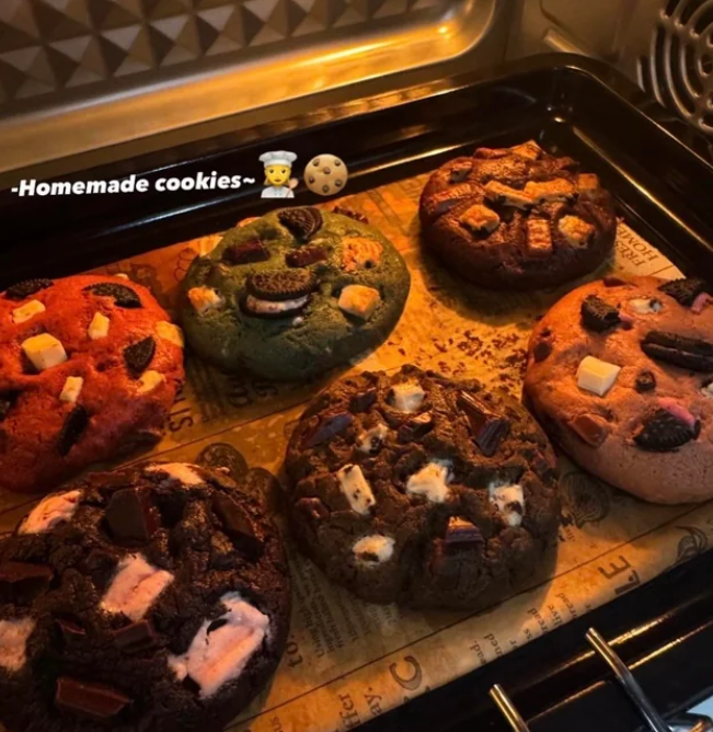 Six large, colorful cookies with various toppings, like chocolate chunks and biscuits, are baking on a tray in an oven. The image features the text "Homemade cookies" with chef and cookie emojis—perfect for anyone who loves cooking from scratch!