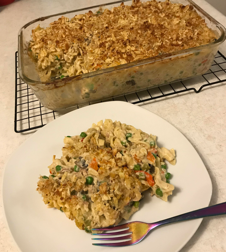 A slice of creamy chicken noodle casserole with vegetables sits on a white plate, next to a baking dish of the same casserole, topped with a crispy golden breadcrumb layer—a comforting classic and perfect for budget meals. A fork rests on the plate.