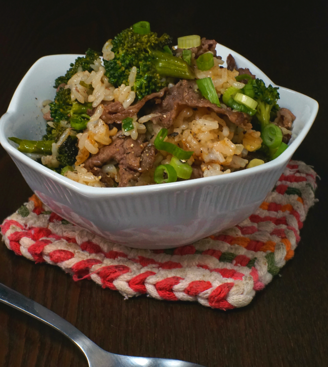 A white bowl filled with beef, broccoli, rice, and chopped green onions sits on a red and white woven mat, offering a delicious budget meal next to a metal spoon on a dark wooden table.