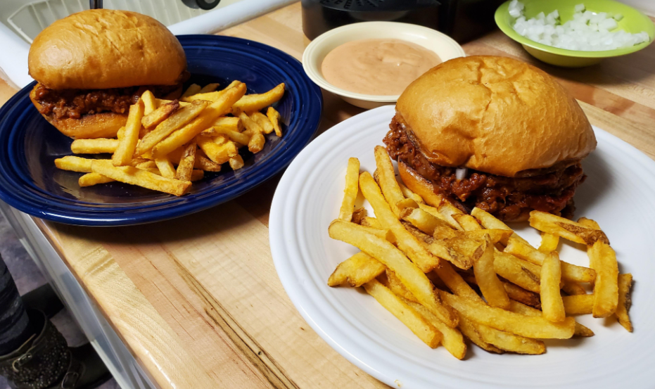 Two plates, each with a barbecue sandwich and French fries, sit on a wooden surface. In the background are bowls of chopped onions and dipping sauce—a delicious option for those seeking satisfying budget meals.